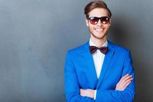 Smile is a part of his style. Cheerful young man in eyewear keeping arms crossed and smiling at camera while standing against grey background photo