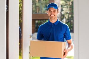 Delivery with smile. Cheerful young courier holding a cardboard box while standing at the entrance of apartment photo