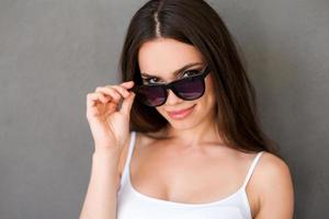 sentirse coqueta. una joven sonriente ajustando las gafas y mirando la cámara mientras se enfrenta a un fondo gris foto