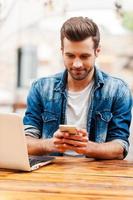 Quick text while working. Handsome young man holding mobile phoneand looking at it while sitting at the wooden table outdoors photo