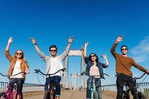 Enjoying fun ride. Low angle view of four young cheerful people riding their bicycles and keeping arms raised photo