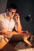 Busy working even at night. Top view of confident young man in eyewear talking on the mobile phone and working on his laptop while sitting at his working place at night time photo