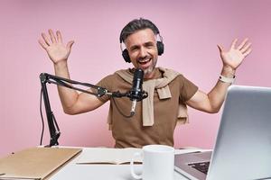 Excited mature man using microphone and gesturing while recording podcast against pink background photo