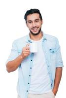 Taking a coffee break. Handsome young Indian man holding coffee cup and smiling while standing against white background photo