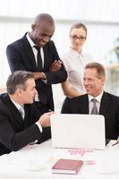 Business team at meeting. Two cheerful mature man in formalwear sitting at the table and communicating while two another people standing close to them photo