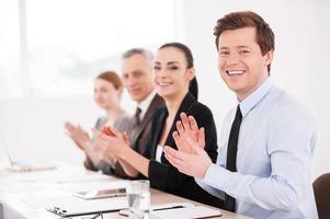 Business seminar. Side view of smiling business people sitting in a row at the table and applauding to you photo