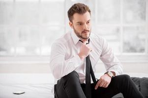 Tired executive. Young man in formalwear taking off his necktie while sitting on the bed photo