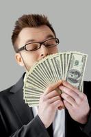 Money smelling. Confident young man in formalwear and glasses holding paper currency and smelling it while standing against grey background photo
