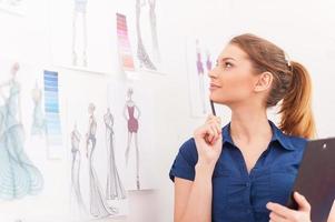 Creating a new fashion look. Beautiful female fashion designer holding clipboard and looking at the sketches hanging on the wall photo