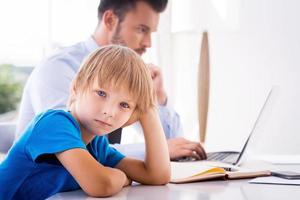 Feeling so lonely. Sad little boy holding hand in hair and looking at camera while his busy father working in the background photo