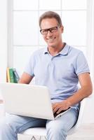 hombre de negocios casual confiado. hombre maduro confiado trabajando en una laptop y sonriendo mientras se sienta en la silla foto