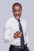 Furious businessman. Furious young African man in shirt and tie holding mobile phone and shouting while standing against grey background photo