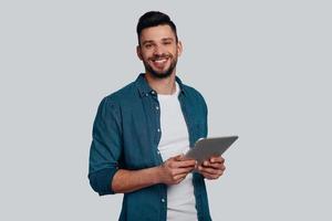 Ready to help. Handsome young man holing digital tablet and looking at camera with smile while standing against grey background photo