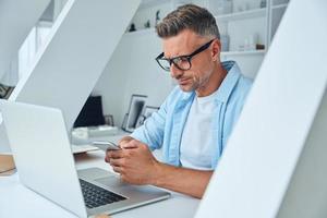 Confident mature man using smart phone while sitting at the office desk photo