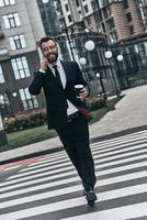 Confident businessman. Full length of young man in full suit talking on the phone and smiling while crossing the street photo