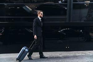 Executive on the go. Full length of young man in full suit pulling luggage while walking outdoors photo