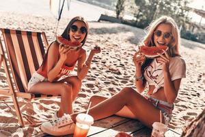 disfrutando de un día cálido. dos atractivas mujeres jóvenes sonriendo y comiendo sandía mientras se sientan en la playa foto