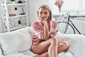 Searching for ideas. Attractive young woman using smart phone and smiling while sitting on bed at home photo