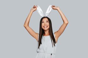 Doing whatever she wants. Beautiful young Asian woman touching her bunny ears and smiling while standing against grey background photo