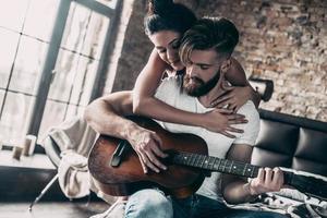 Guitar fun. Handsome young bearded man sitting in bed at home and playing guitar while attractive woman embracing him photo