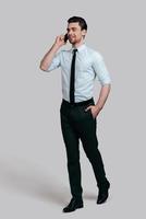 Pleasant business talk. Full length of handsome young man talking on smart phone and keeping hand in pocket while standing against grey background photo