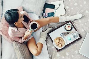 Top view of beautiful young woman in pajamas enjoying hot drink while resting in bed at home photo