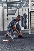 preparándose para el entrenamiento. un apuesto joven africano con ropa deportiva atándose los zapatos mientras hace ejercicio en el gimnasio foto