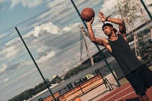 disparo cuidadoso. joven africano vestido con ropa deportiva lanzando una pelota mientras juega baloncesto al aire libre foto