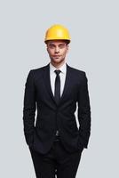 Safety comes first. Good looking young man in hardhat keeping hands in pockets and looking at camera while standing against grey background photo