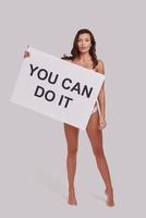 I believe in you  Full length of attractive young woman covering with a poster and looking at camera with a smile while standing against grey background photo