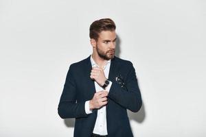 Confident in his style. Handsome young man in full suit adjusting his sleeve and looking away while standing against grey background photo