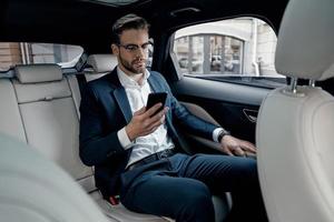 joven perfeccionista. un joven apuesto con traje completo mirando su teléfono inteligente mientras está sentado en el auto foto