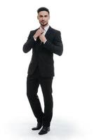 Looking just perfect. Full length of handsome young man in full suit adjusting his necktie and looking at camera while standing against white background photo