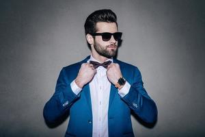 Mr. Perfection. Close-up of handsome young man wearing sunglasses adjusting his bow tie and looking away while standing against grey background photo