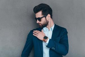 Shake it off. Close-up of young handsome man in sunglasses shaking off invisible dust from his shoulder while standing against grey background photo