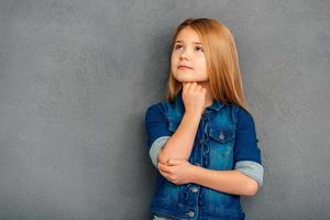 tal vez esta sea una buena idea hermosa niña sosteniendo la mano en la barbilla y mirando hacia otro lado mientras está de pie contra un fondo gris foto