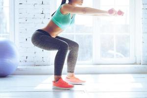 ardor en las nalgas. vista lateral de una mujer joven en ropa deportiva haciendo sentadillas y sosteniendo pesas mientras está de pie frente a la ventana en el gimnasio foto