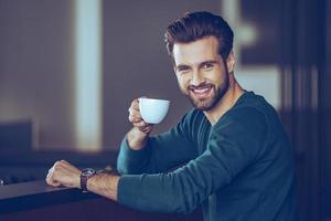 Salute Side view of handsome young man holding coffee cup and looking at camera with smile while sitting at bar counter photo