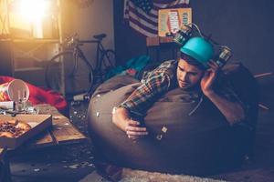 apuesto joven con sombrero de cerveza usando su teléfono inteligente mientras está acostado en una bolsa de frijoles en una habitación desordenada después de la fiesta foto