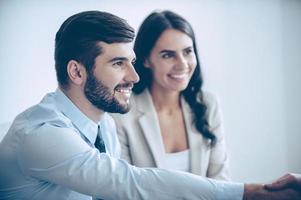 es un placer trabajar contigo parte de primer plano de jóvenes apuestos que se dan la mano con una sonrisa mientras están sentados en la oficina con sus compañeros de trabajo foto