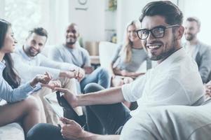 Confident psychologist. Group of young cheerful people sitting in circle and discussing something while young man holding digital tablet and looking over shoulder with smile photo