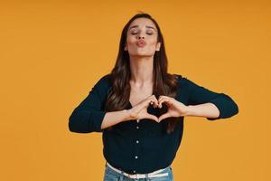 Attractive young woman in casual clothing making heart gesture while standing against yellow background photo
