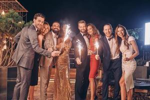 Group of beautiful people in formalwear holding sparklers and smiling while spending time on outdoor party photo