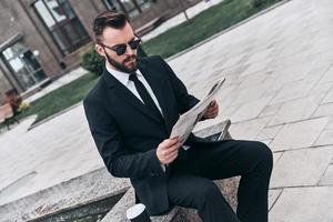 Interesting article. Good looking young man in full suit reading a newspaper while sitting outdoors photo
