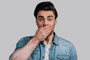 Keeping your secret. Good looking young man in casual wear covering mouth with hand and looking at camera while standing against grey background photo