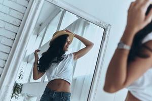 belleza juguetona. reflejo de una joven atractiva probándose el sombrero para el sol mientras pasa tiempo en casa foto