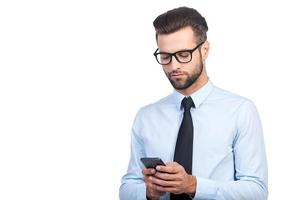 Typing business message. Confident young handsome man in shirt and tie holding mobile phone and looking at it while standing against white background photo