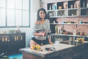 mirando mi día con café recién hecho. Hermosa joven mujer de raza mixta sosteniendo un frasco con café y sonriendo mientras está de pie en la cocina en casa foto