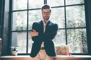 Confident and successful. Confident young man in smart casual wear keeping arms crossed and looking at camera while standing in front of the big window in office or cafe photo