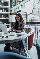 Her job is her hobby. Beautiful young woman using laptop and smiling while sitting in restaurant photo